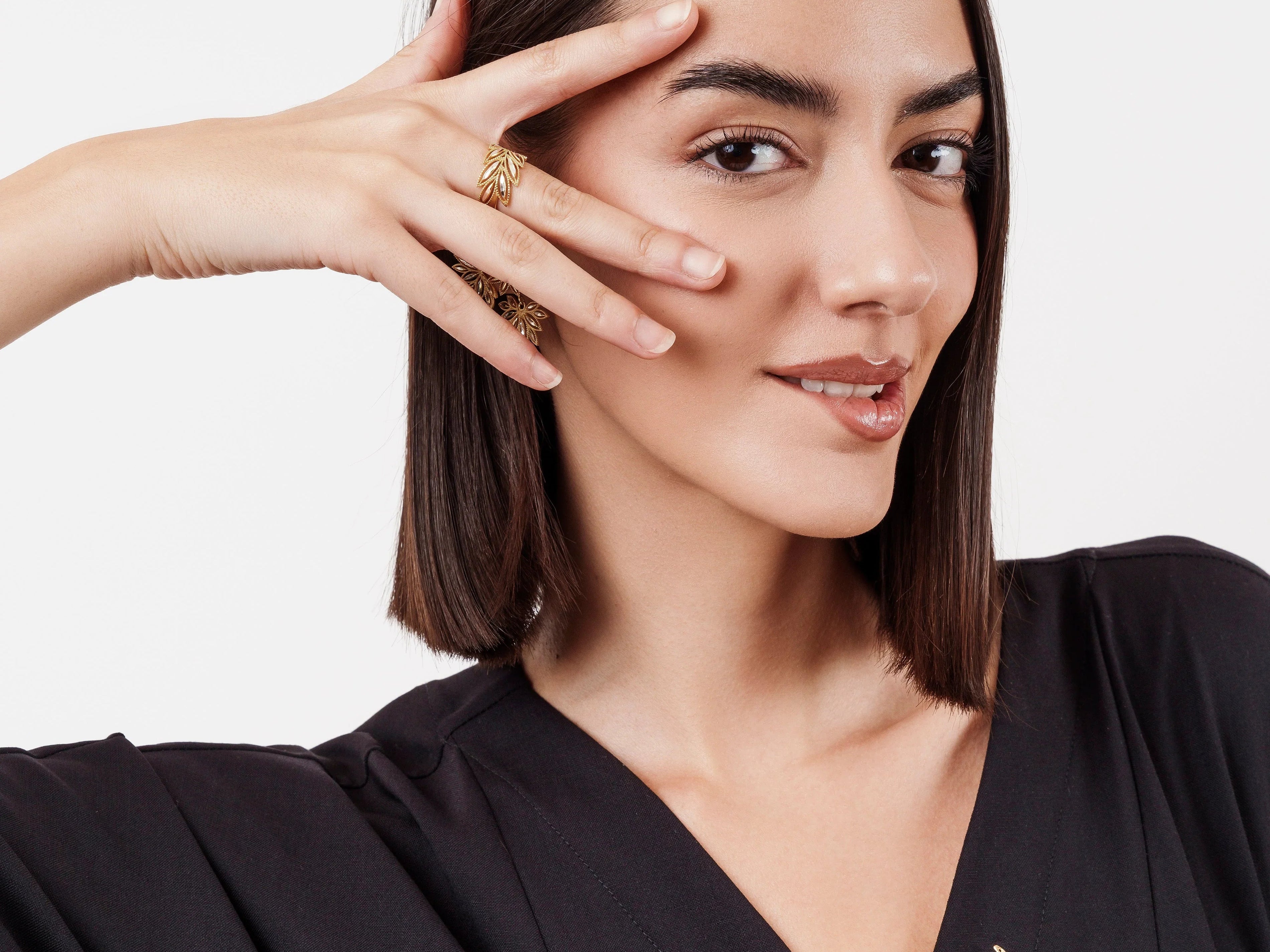 Smiling woman with straight dark hair wearing black V-neck top and gold leaf jewelry, posing with hand near face
