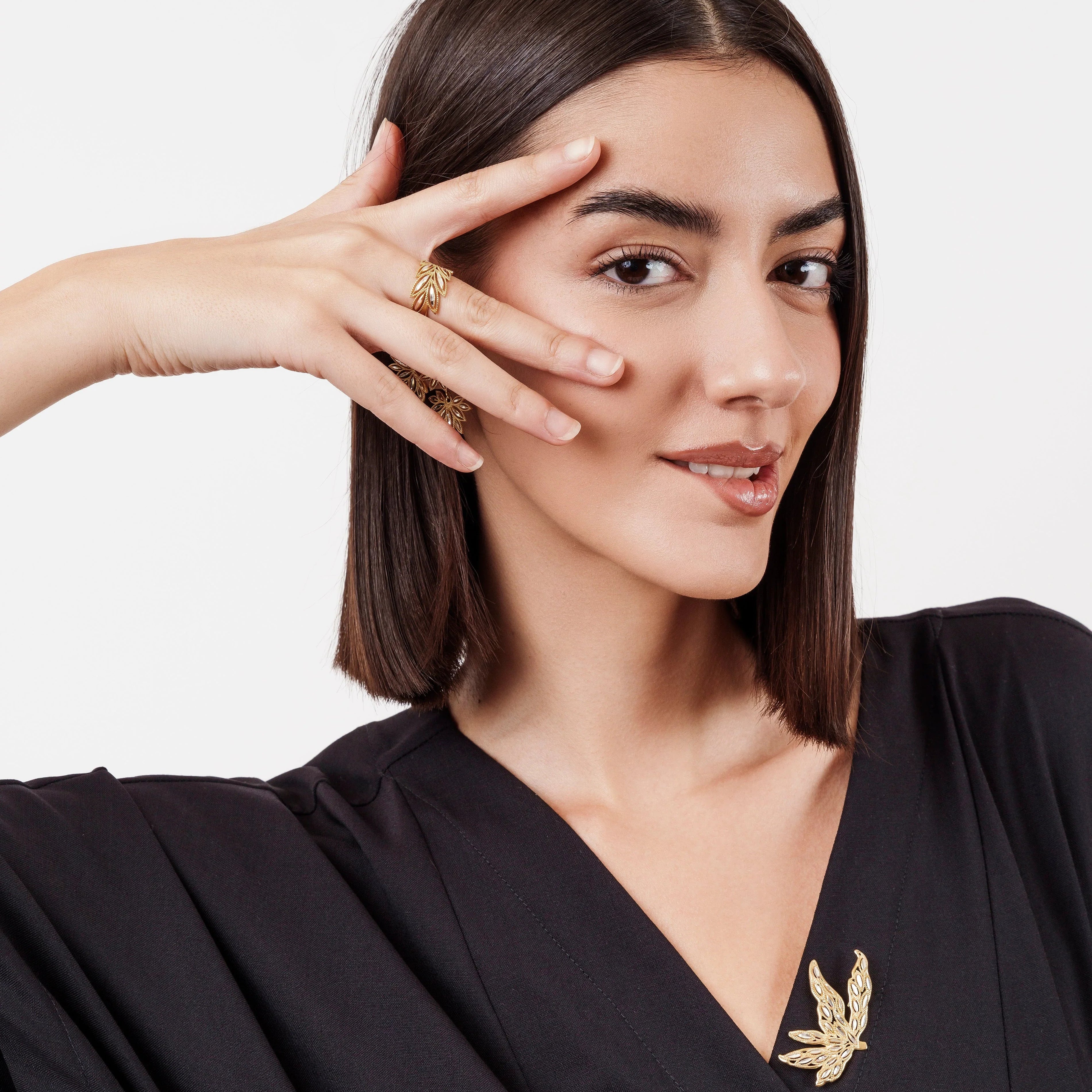 Smiling woman with straight dark hair wearing black V-neck top and gold leaf jewelry, posing with hand near face