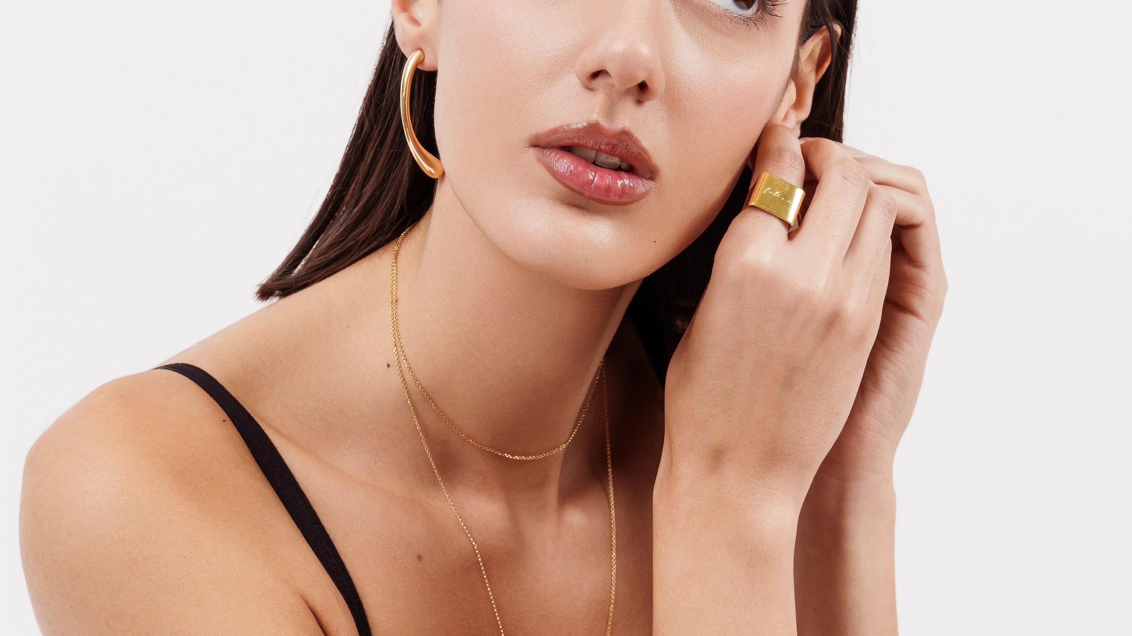 Portrait of woman wearing gold hoop earrings, layered gold necklaces, and gold statement ring with black tank top