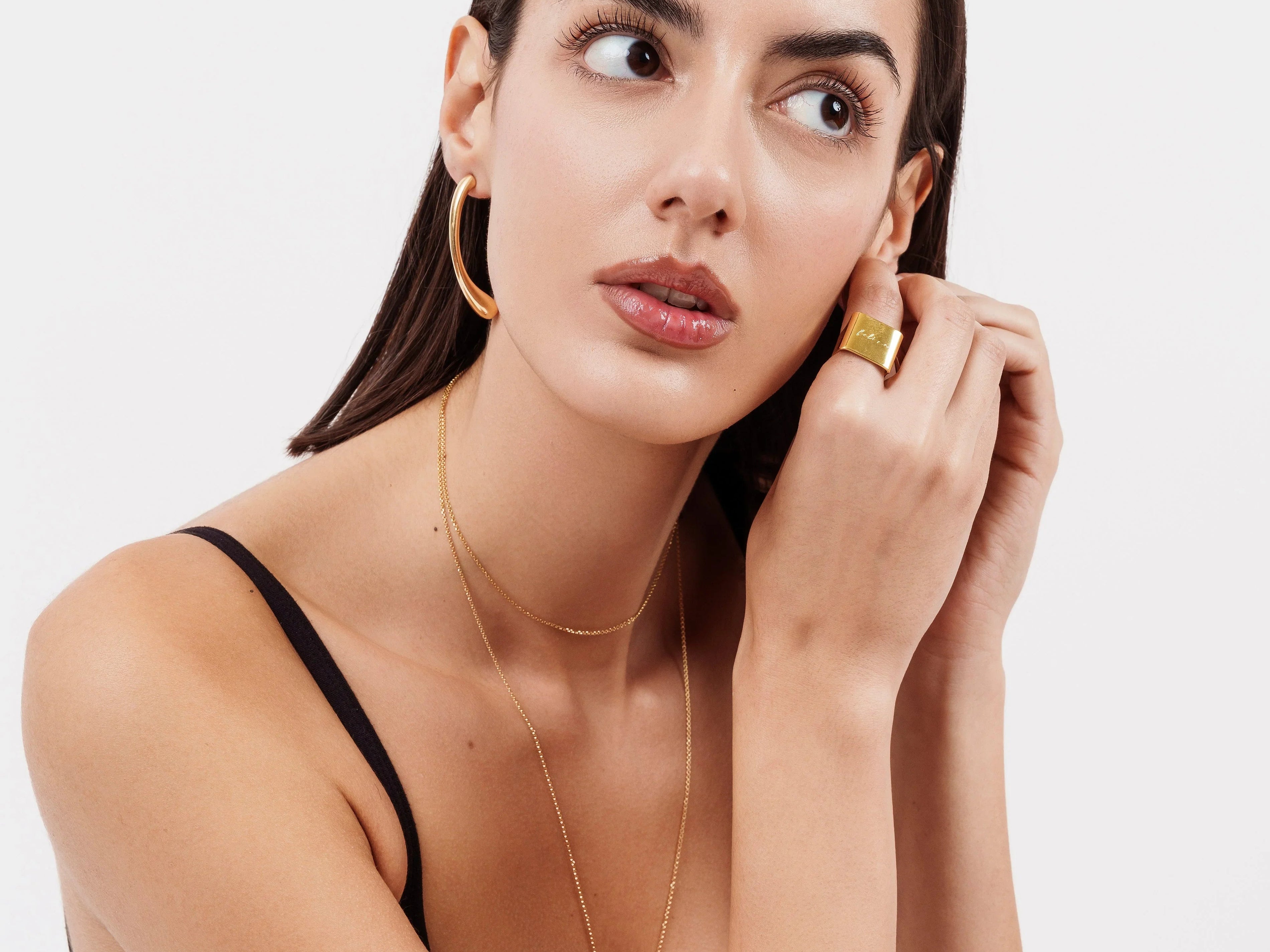 Portrait of woman wearing gold hoop earrings, layered gold necklaces, and gold statement ring with black tank top
