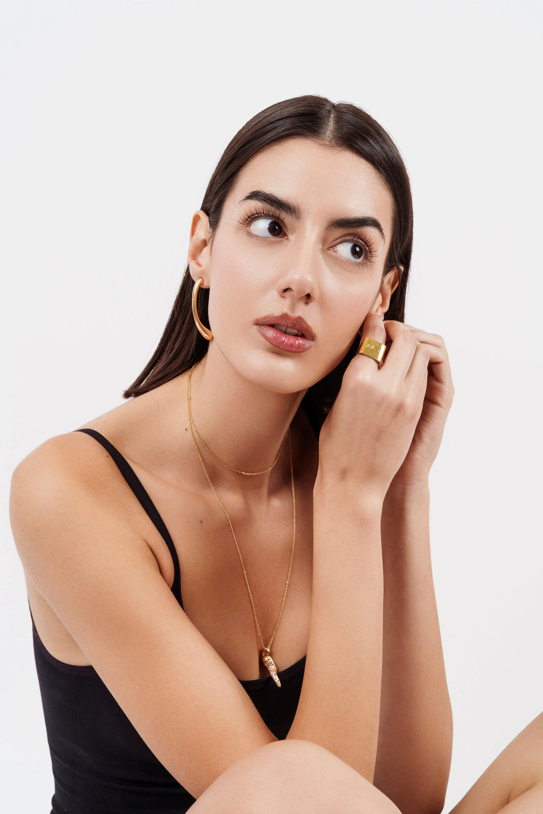 Portrait of woman wearing gold hoop earrings, layered gold necklaces, and gold statement ring with black tank top