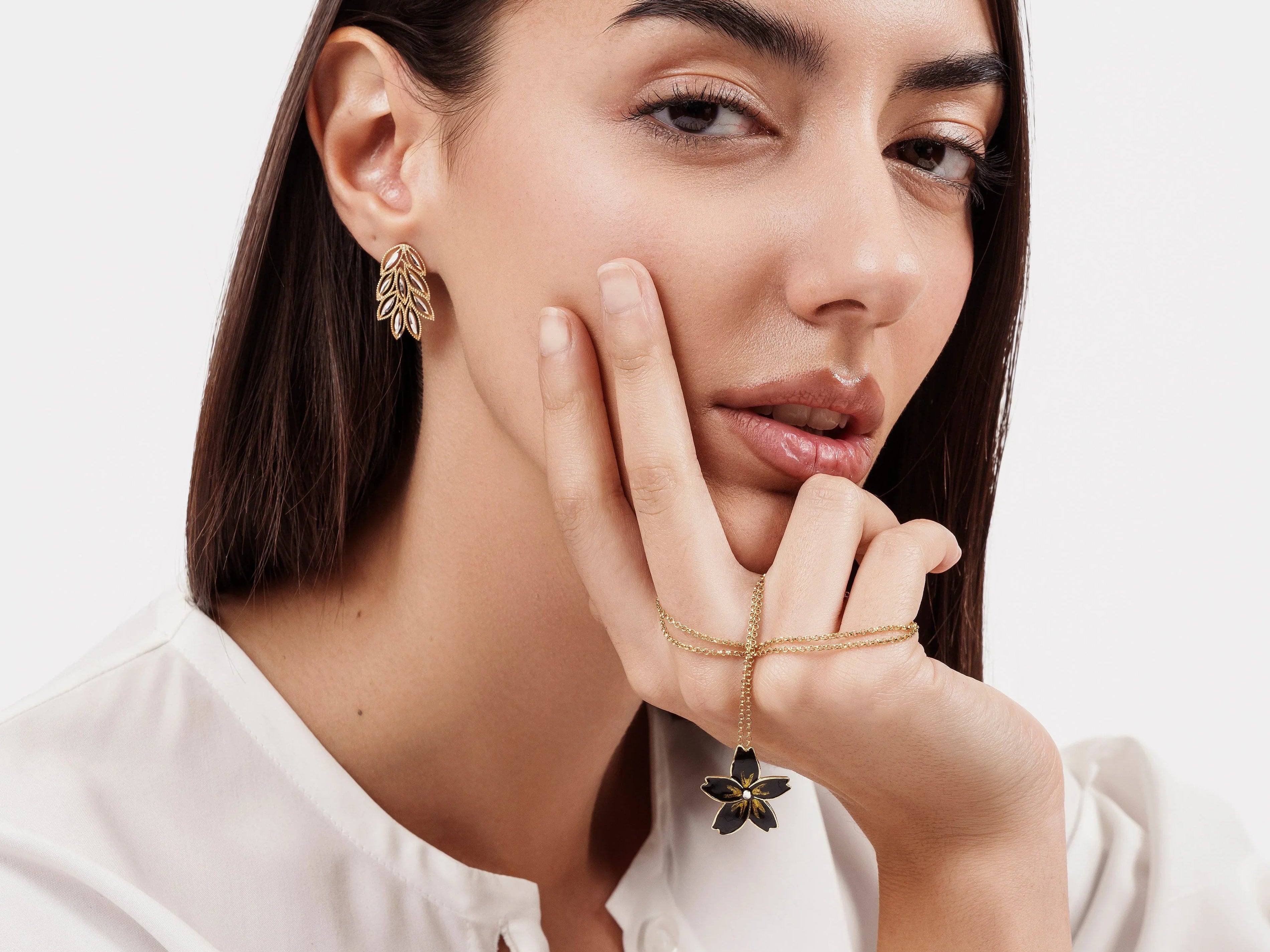 Close-up of woman with dark hair wearing gold leaf earrings and holding a gold flower pendant necklace