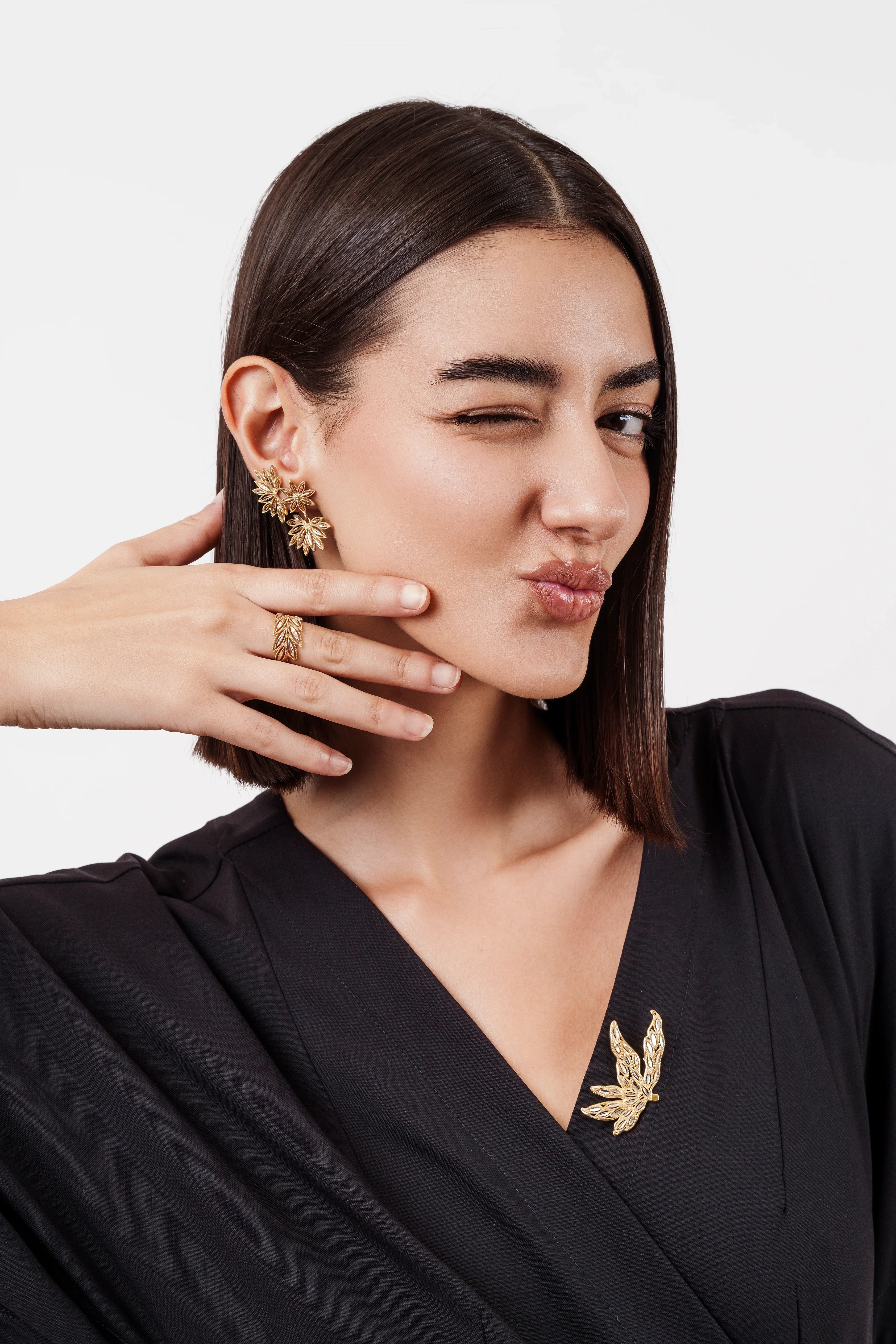 Woman in black top wearing gold floral earrings, matching ring, and leaf-shaped brooch, winking