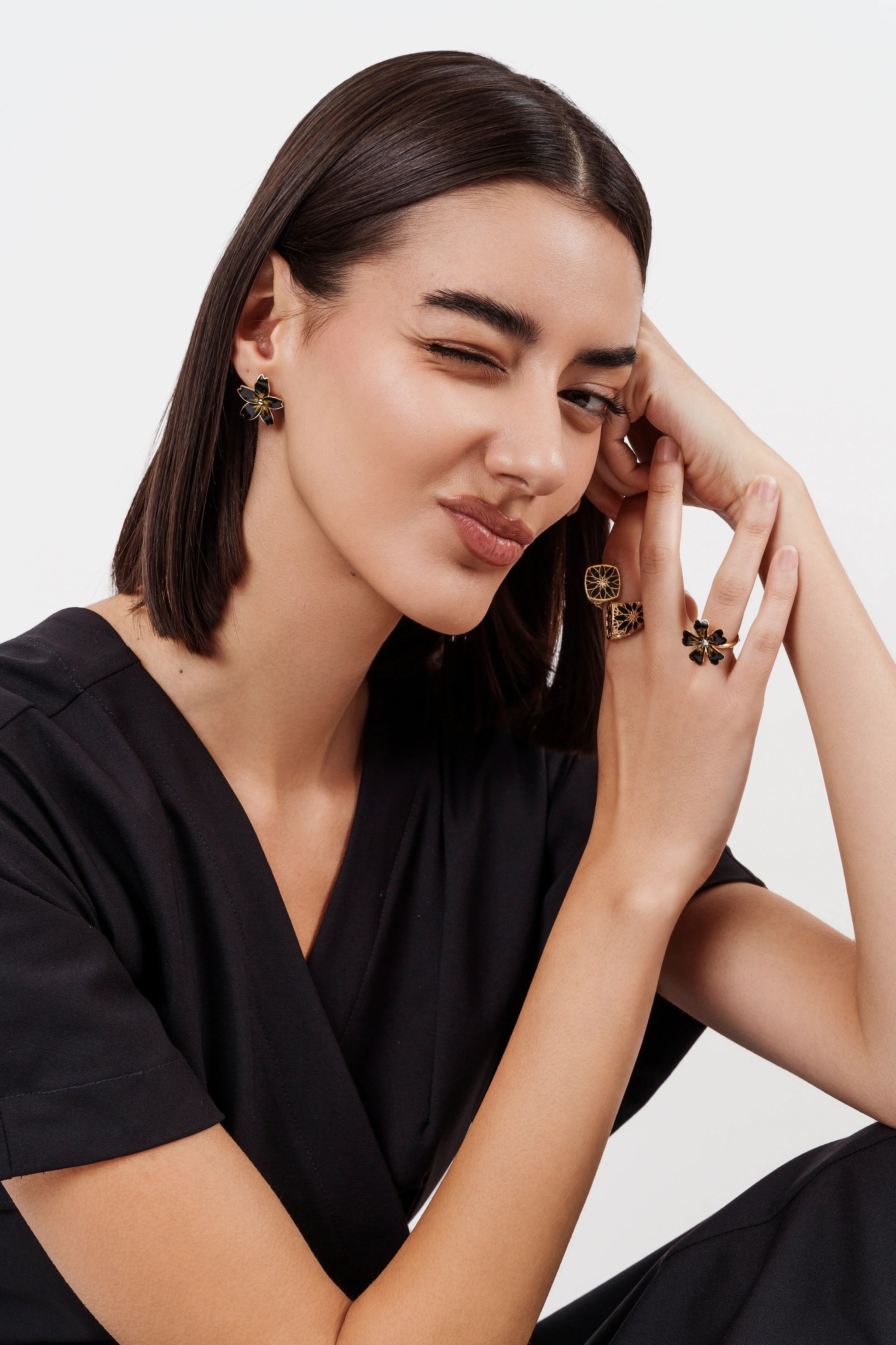 Woman with short dark hair wearing black outfit, winking and showcasing gold black floral earrings and rings