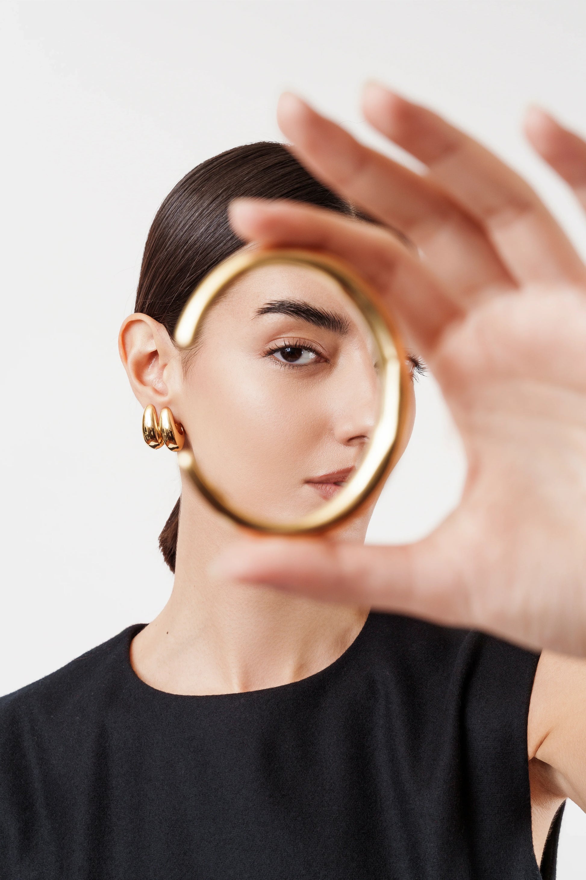 Woman with dark hair in black top holding gold bangle bracelet near face, wearing gold hoop earrings