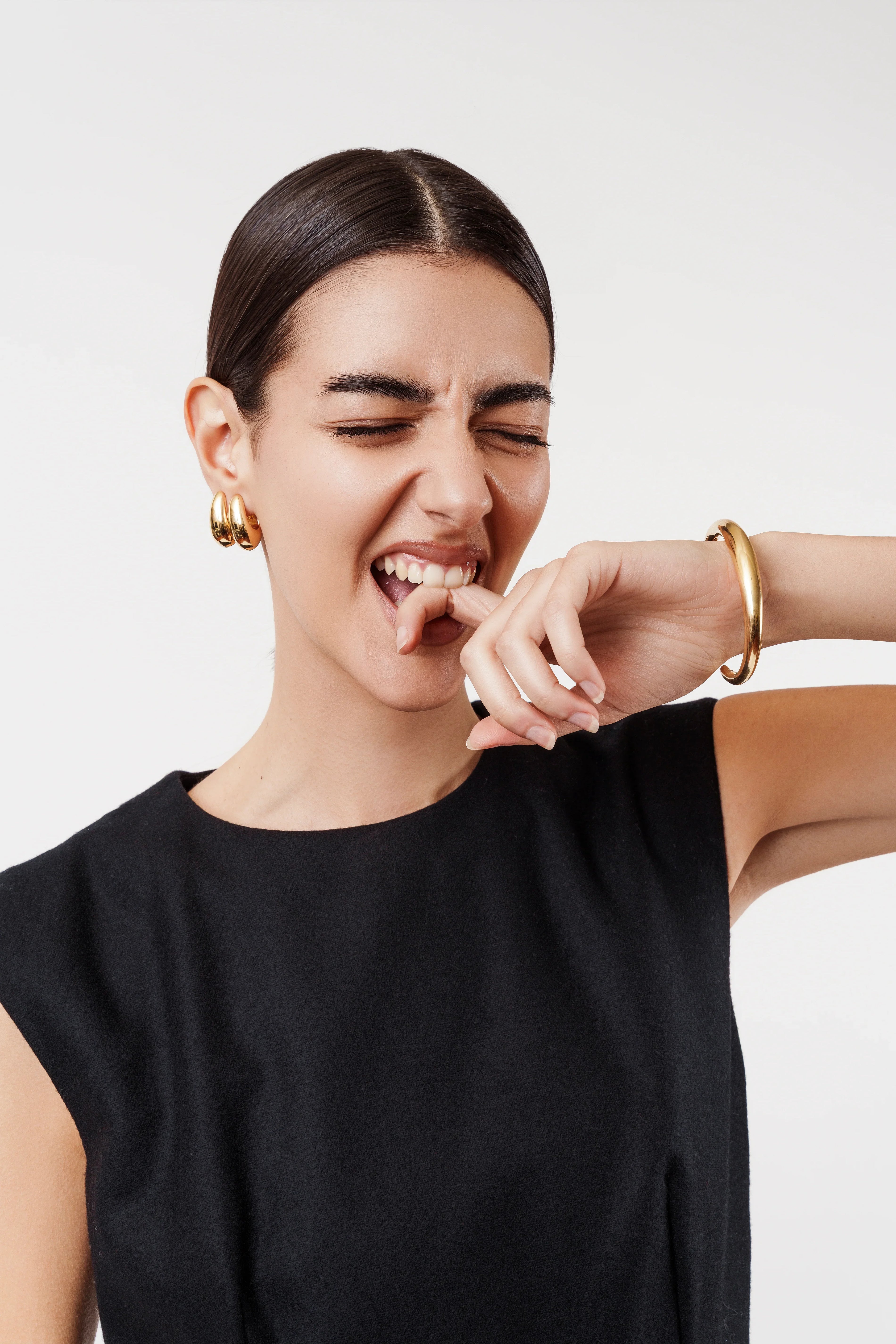Woman in black sleeveless top wearing gold hoop earrings and bracelet, biting finger playfully