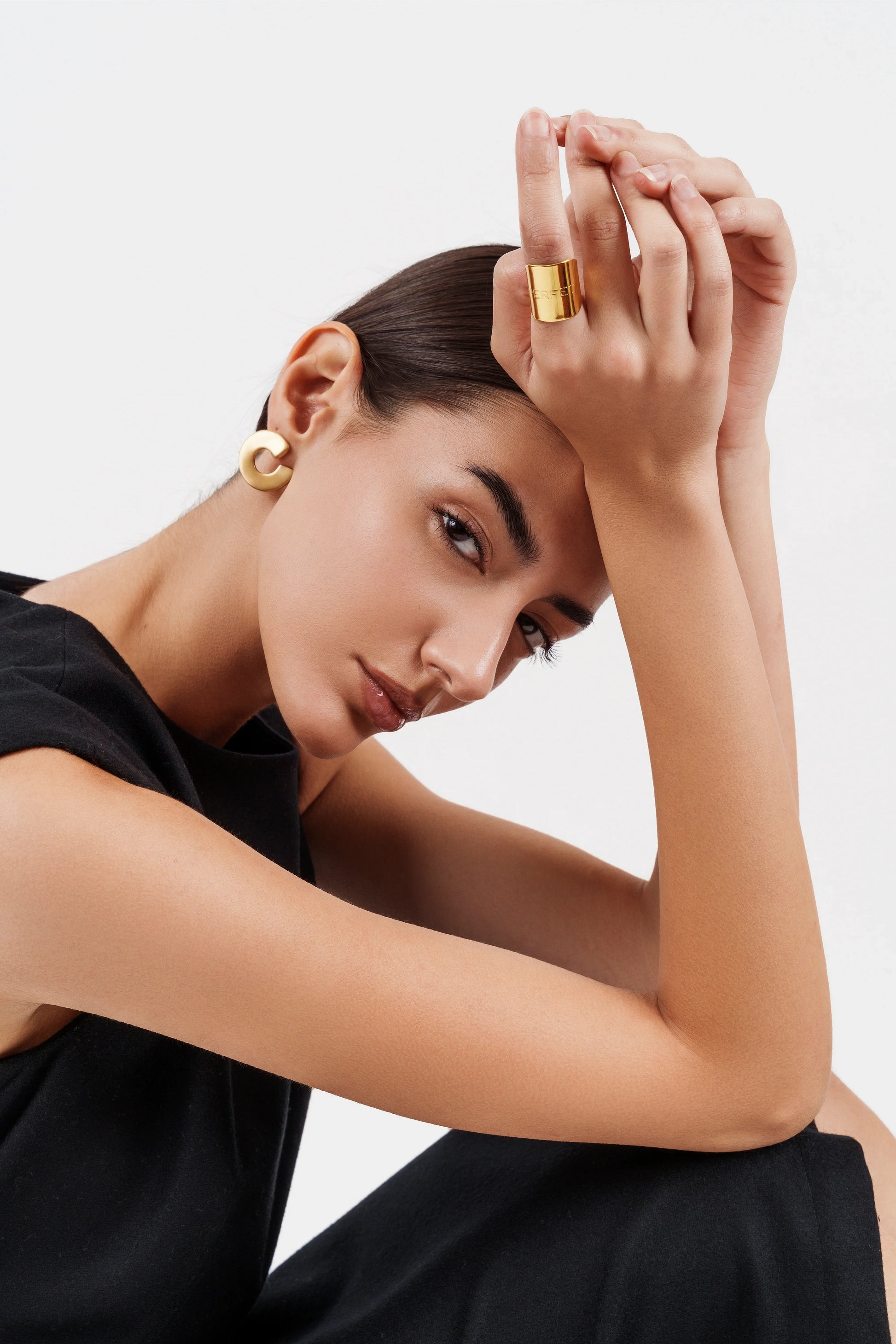 Portrait of woman in black dress wearing gold hoop earrings and wide gold ring against white background