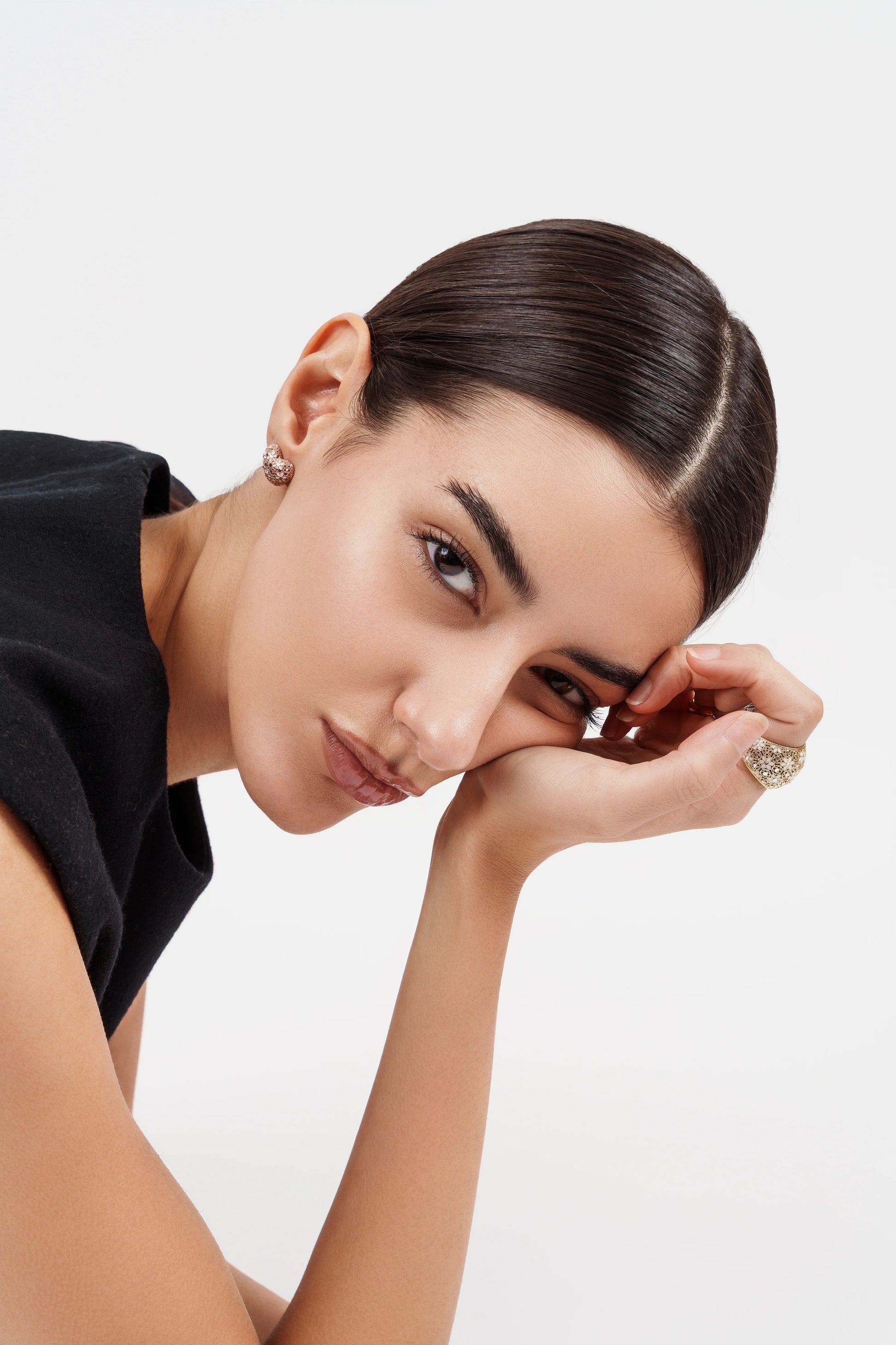 Close-up portrait of woman with sleek hair, wearing textured gold earrings and a gold ring, black top