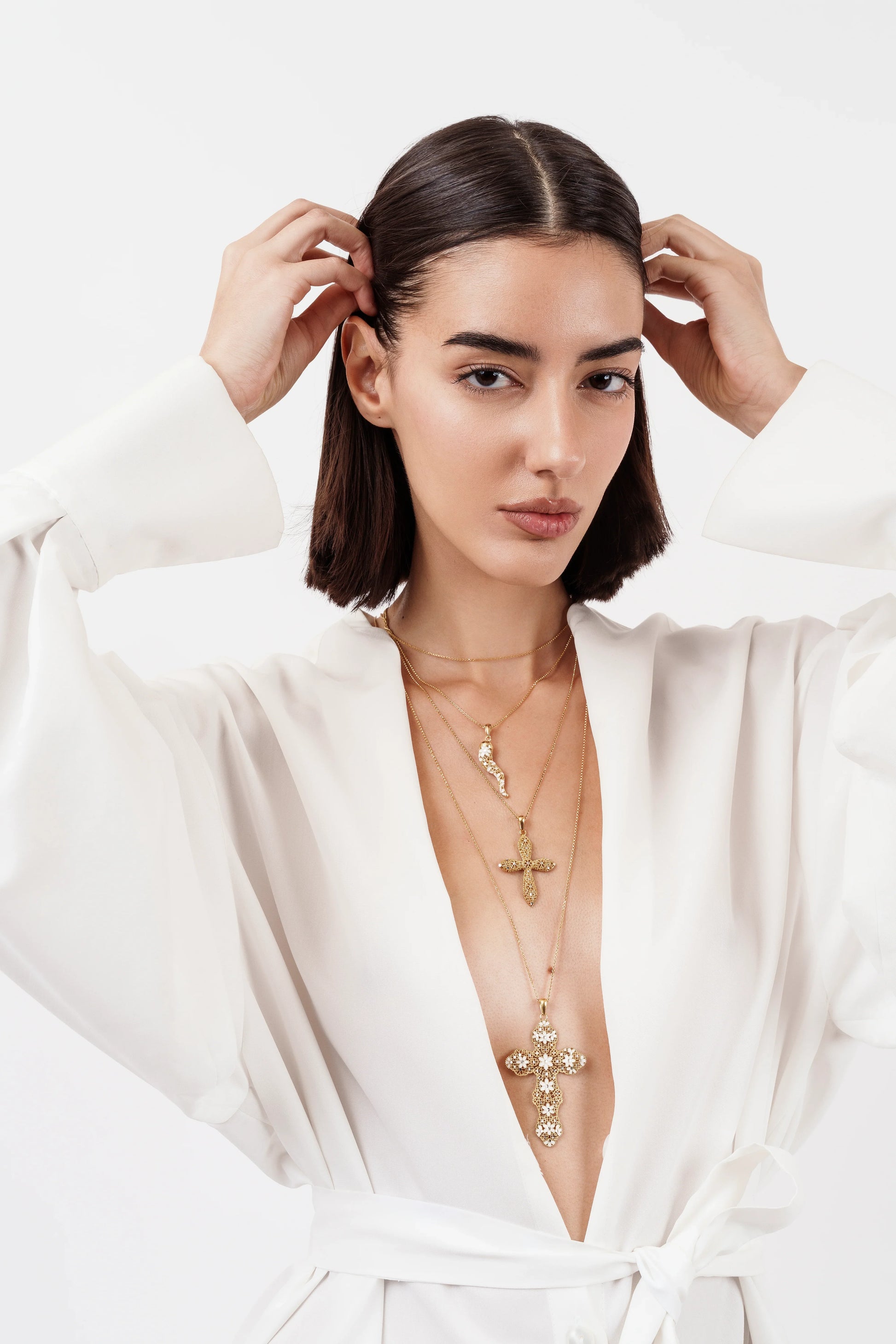 Woman in white robe wearing layered gold cross necklaces with floral details against white background