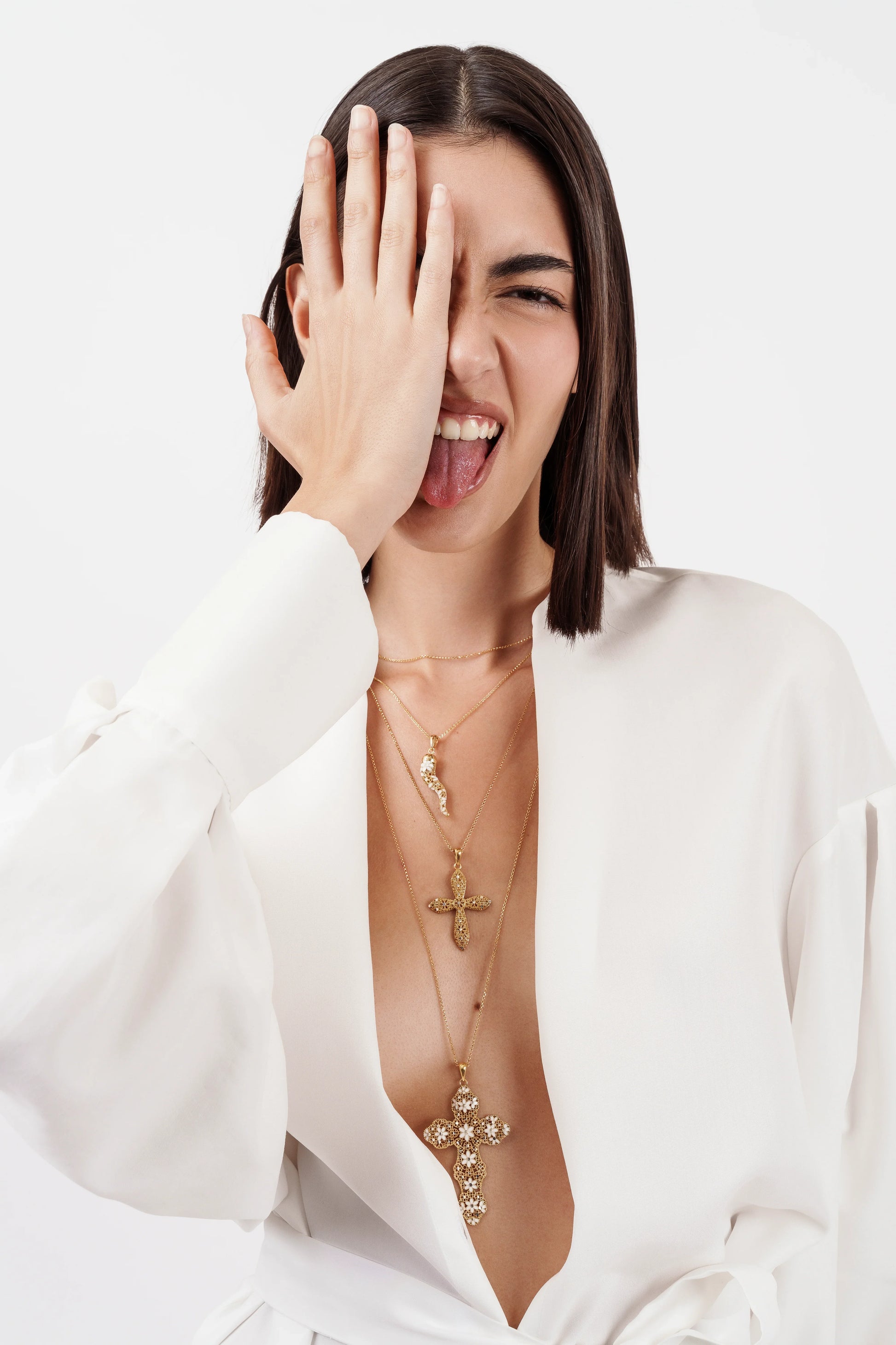 Woman in white blouse wearing layered gold cross necklaces, covering one eye, sticking out tongue