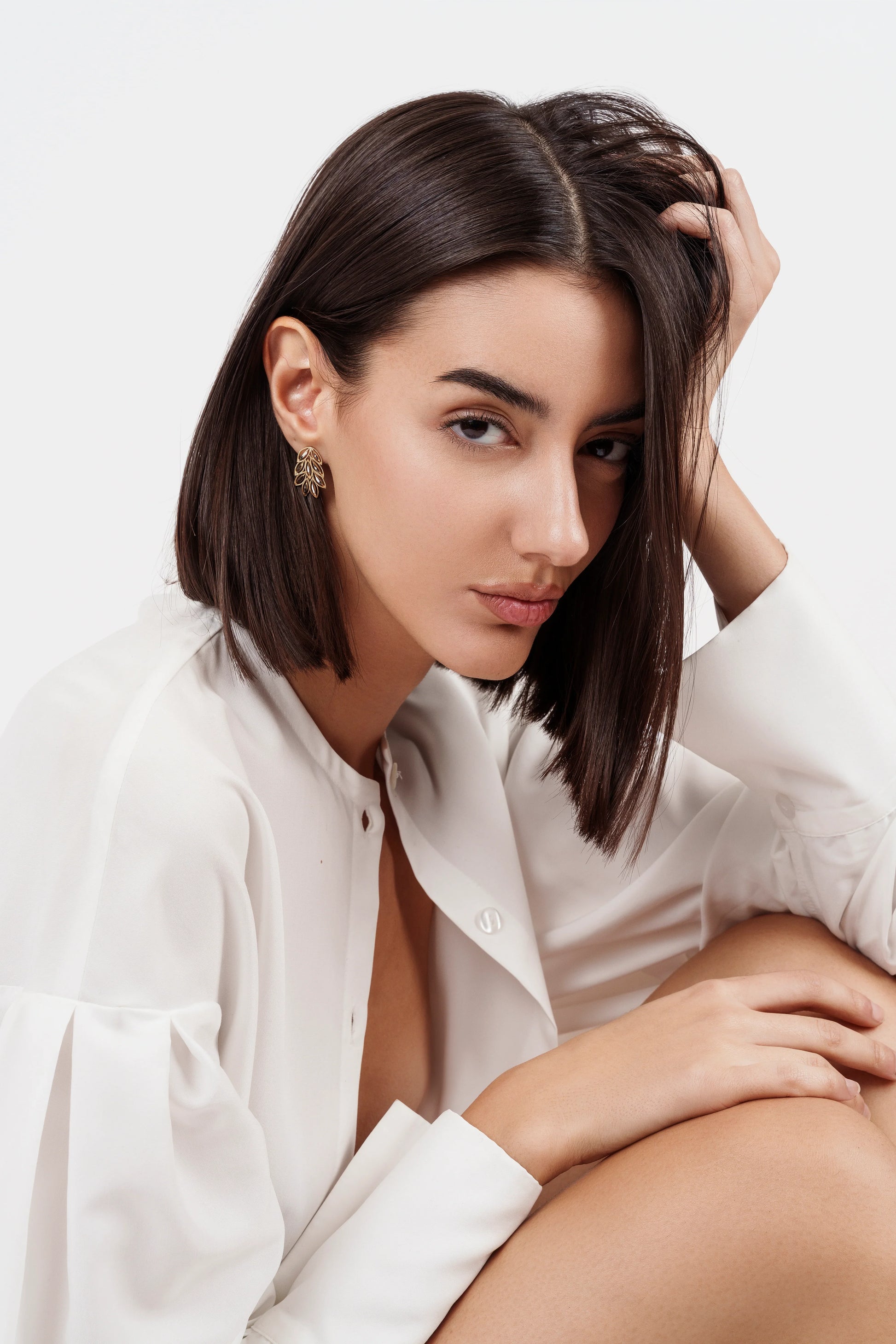 Close-up portrait of woman with short dark hair wearing gold leaf earrings and white blouse on white background