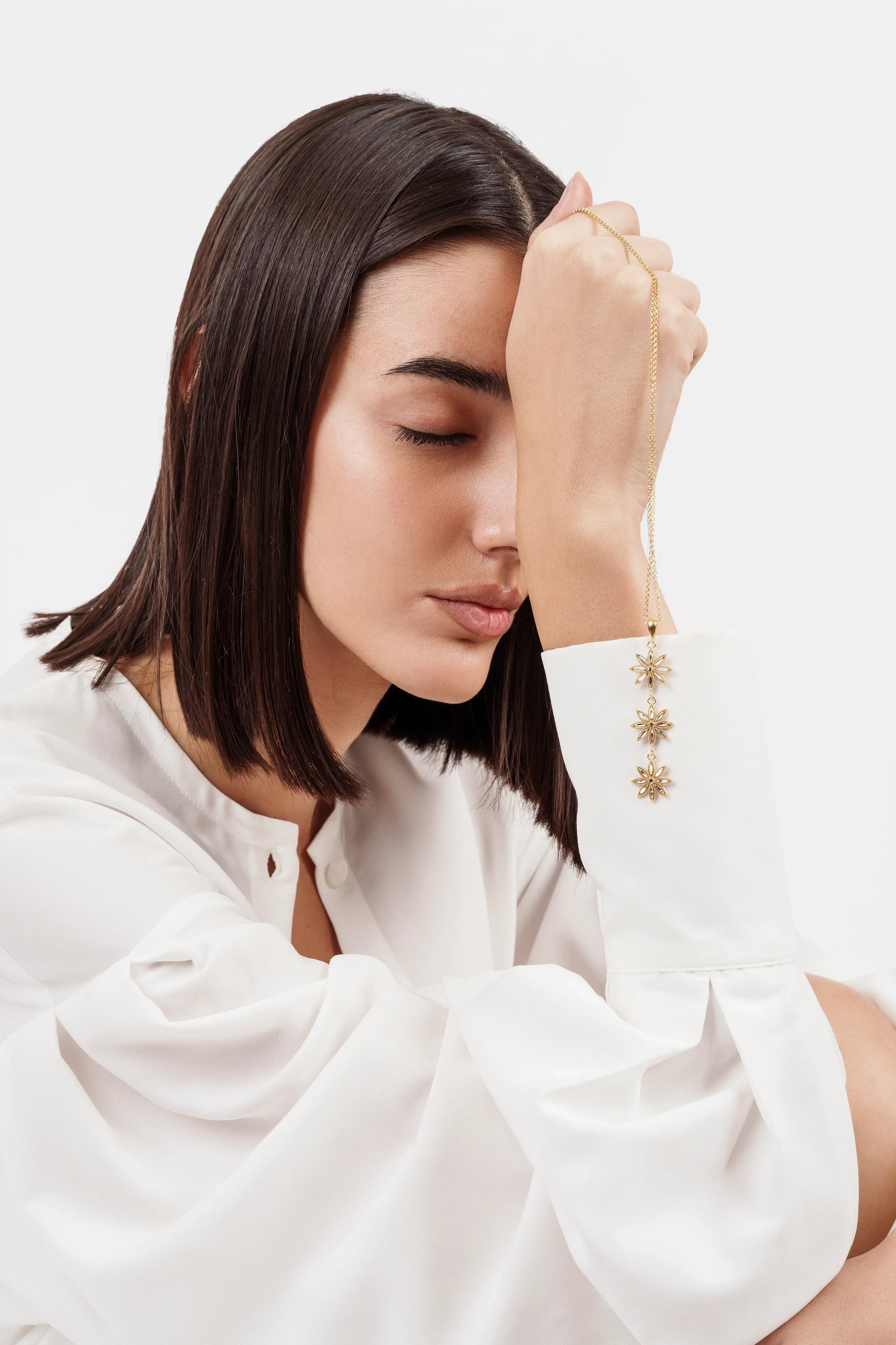 Woman with straight dark hair in white blouse holding gold floral pendant necklace with closed eyes