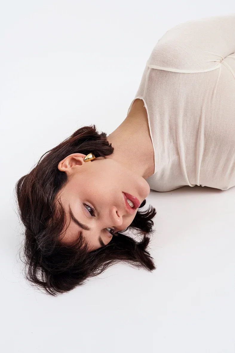 Woman lying down with short dark hair, wearing gold earrings and a beige top on white background