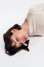 Woman lying down with short dark hair, wearing gold earrings and a beige top on white background